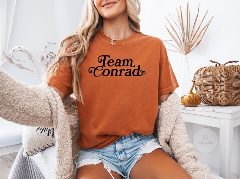 May include: An orange t-shirt with the text "Team Conrad" in a black script font. The shirt is worn with denim shorts and a cream-colored cardigan. A decorative pumpkin and woven basket are in the background.
