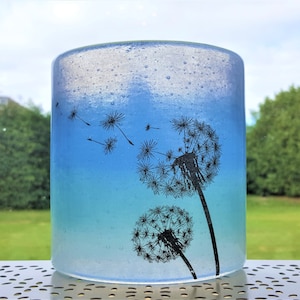 May include: A clear glass cylinder vase with a blue to white ombre effect. The vase has a black silhouette of dandelions with seeds blowing in the wind.