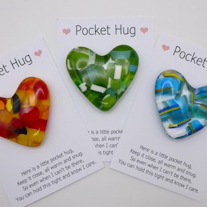 Op de afbeelding: Drie hartvormige glazen 'Pocket Hugs' in rood, groen en blauw, elk met een uniek mozaïekontwerp. Elk hart ligt op een kaart met de tekst 'Pocket Hug' en een boodschap van troost.