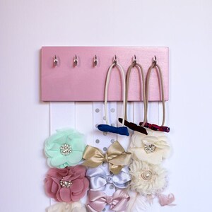 May include: Pink wooden headband holder with hooks and white ribbon strips with various fabric flowers and bows attached.