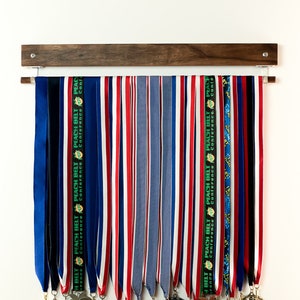 May include: A wooden wall-mounted medal display rack with a row of blue, red, white, and black ribbons with medals hanging from the bottom. The medals are gold, silver, and bronze and have various designs.