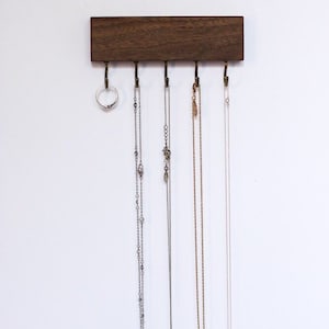 May include: A wooden wall-mounted jewelry organizer with five hooks. A ring and four necklaces are hanging from the hooks. The necklaces are silver, gold, and a combination of silver and gold.