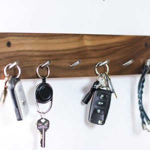May include: A wooden key rack with five hooks, holding a set of keys, a retractable key chain, a car key, and a pair of sunglasses.