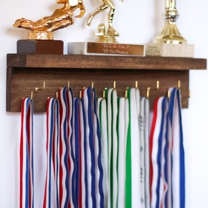 May include: A wooden wall-mounted medal display with hooks for hanging ribbons. The display has a shelf on top with two gold trophies and a plaque that reads "U.S.A. Elite 7 71st Season".