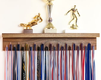 May include: A wooden wall-mounted medal display with hooks for hanging ribbons. The display has a shelf on top with two gold trophies and a gold figurine. The ribbons are red, white, blue, and black.