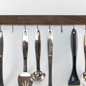 May include: A wooden wall-mounted rack with eight hooks, holding various kitchen utensils, including a spatula, ladle, spoon, and a black silicone brush.