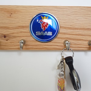 Könnte beinhalten: Ein Schlüsselhalter aus Holz mit vier Haken und einem blauen und silbernen Saab-Logo. Das Logo zeigt einen roten Greif mit einer goldenen Krone.