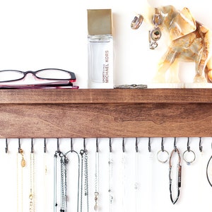 May include: A wooden jewelry organizer with hooks for necklaces and bracelets. The organizer has a shelf on top for storing other items, such as glasses or perfume. The shelf is decorated with a small elephant figurine and a bottle of perfume labeled "Sporty Chic Michael Kors".