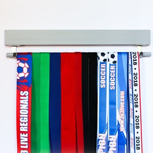 May include: A gray wooden medal display rack with hooks hanging from the bottom.  The rack is holding a variety of colorful ribbons with text that includes "3 Live Regionals", "Soccer", "www.3v3live.com", and "2018".