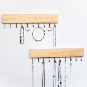 May include: Two wooden wall mounted jewelry organizers with hooks. The top organizer has a silver necklace, a silver bracelet, and two rings hanging from the hooks. The bottom organizer has seven necklaces hanging from the hooks.