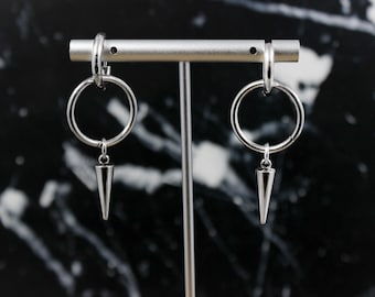 May include: A pair of silver hoop earrings with a dangling spike charm. The earrings are displayed on a stand against a black marble background.