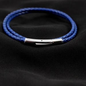 May include: A blue braided leather bracelet with a silver clasp. The bracelet is made of two strands of blue leather and is set against a black background. The clasp is a shiny silver color.