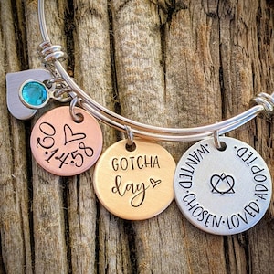 May include: A silver expandable bangle bracelet with three charms. The charms are round and have different engravings. One charm is copper and says "0.3.14.58" with a heart. Another charm is gold and says "GOTCHA day" with a heart. The third charm is silver and says "WANTED. CHOSEN. LOVED. ADOPTED." with a heart and a symbol.