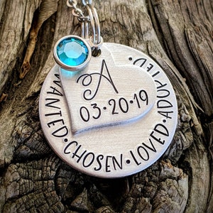 May include: Silver necklace with a heart charm engraved with the letter A, the date 03-20-19, and the words "wanted, chosen, loved, adopted". A small turquoise gemstone dangles from the chain.