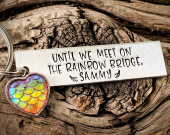 May include: A silver keychain with a heart-shaped charm featuring iridescent rainbow scales. The keychain has a tag that reads "Until we meet on the rainbow bridge. Sammy".