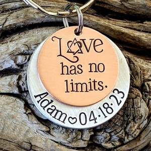 May include: A silver and copper keychain with the words "Love has no limits" and "Adam 04.18.23" engraved on it. The keychain features a heart symbol and a Star of David.