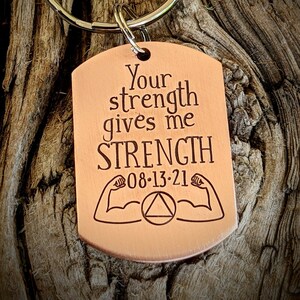May include: A copper-colored dog tag keychain with the words "Your strength gives me STRENGTH" and the date "08-13-21" engraved. The tag also features illustrations of flexing arms and a triangle symbol.