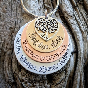May include: A silver, gold, and copper layered keychain with a heart-shaped charm featuring a tree design. The keychain has the text "Gotcha day Braxton 03.15.24 Wanted. Chosen. Loved. Follow..."