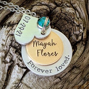 May include: A silver necklace with multiple pendants. The top pendant is heart-shaped with the date "07.29.21" engraved. Below, a round pendant is inscribed with "Mayah Flores" and "Forever loved.". A teal gemstone is set at the top.