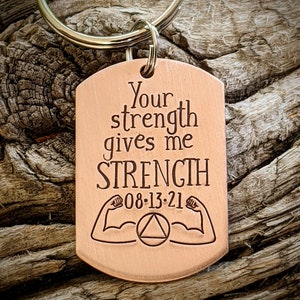 May include: A copper-colored dog tag keychain with the engraved text "Your strength gives me STRENGTH" and the date "08-13-21". The tag features illustrations of flexing arms and a triangle within a circle. The keychain has a silver ring.