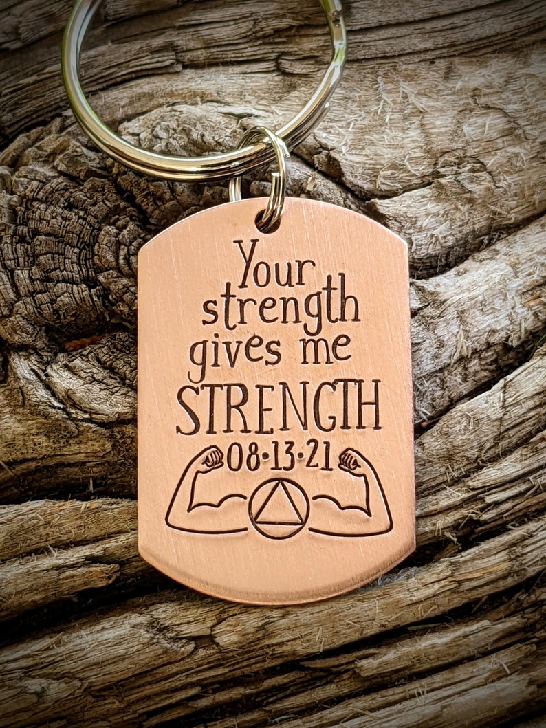 May include: A copper-colored dog tag keychain with the engraved text "Your strength gives me STRENGTH" and the date "08-13-21". The tag features illustrations of flexing arms and a triangle within a circle. The keychain has a silver ring.