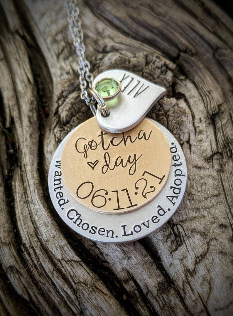 May include: Silver necklace with a heart charm engraved with "ALLY" and a round charm with the text "Gotcha day 06.11.21 Wanted. Chosen. Loved. Adopted."