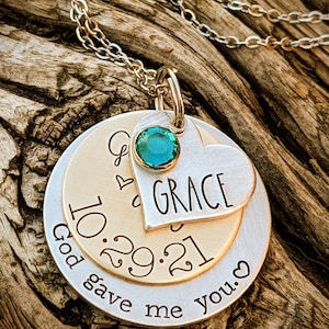 May include: A silver necklace with a layered pendant. The top layer is a heart with the word "GRACE" engraved on it. The middle layer is a round disc with the date "10.29.21" engraved on it. The bottom layer is a round disc with the text "God gave me you." engraved on it. A teal gemstone is attached to the top of the pendant.