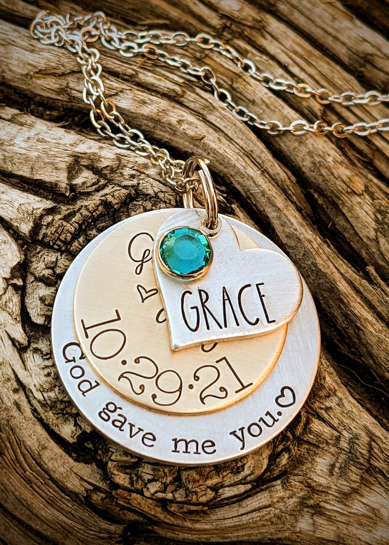 May include: A silver necklace with a layered pendant. The top layer is a heart with the word "GRACE" engraved on it. The middle layer is a round disc with the date "10.29.21" engraved on it. The bottom layer is a round disc with the text "God gave me you." engraved on it. A teal gemstone is attached to the top of the pendant.