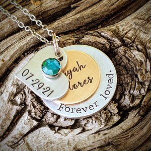 May include: A layered pendant necklace with a silver chain. The pendant features a heart-shaped tag with "07.29.21" and a teal gemstone. Below, a gold-toned disc is inscribed with a name, and a silver disc reads "Forever loved."