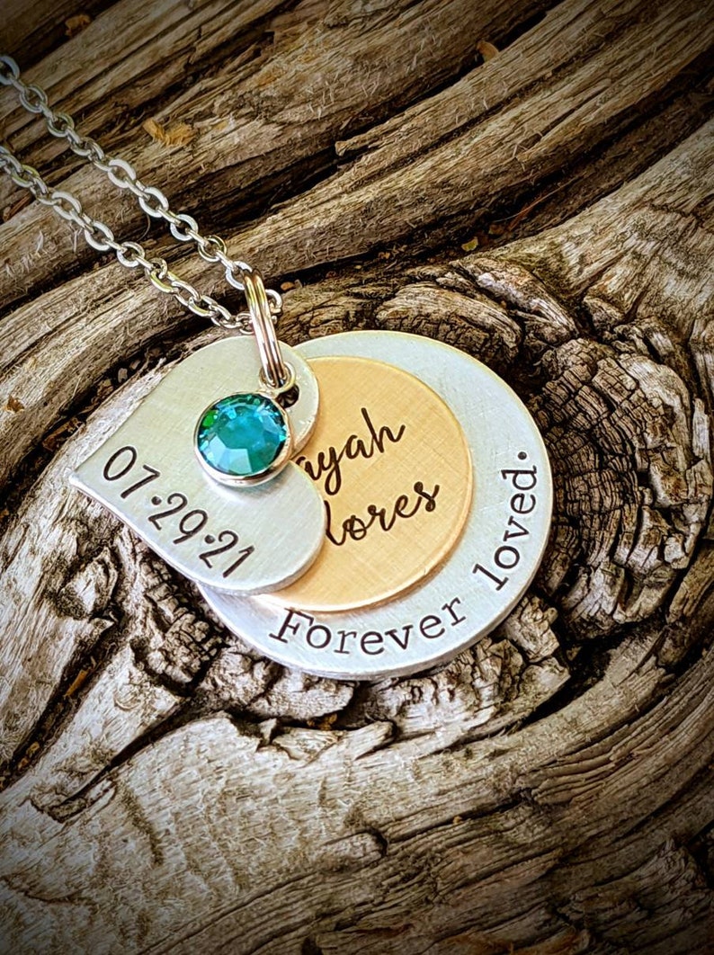 May include: A layered pendant necklace with a silver chain. The pendant features a heart-shaped tag with "07.29.21" and a teal gemstone. Below, a gold-toned disc is inscribed with a name, and a silver disc reads "Forever loved."