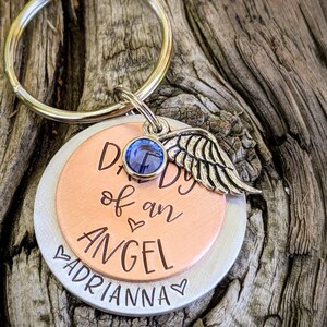 May include: A silver and copper keychain with a blue rhinestone and a silver angel wing charm. The keychain has the text "Daddy of an Angel" and "Adrianna" engraved on it.