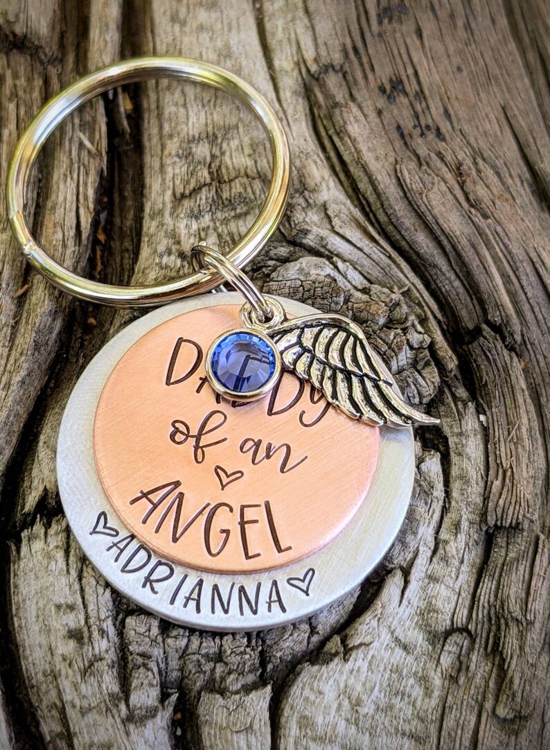 May include: A silver and copper keychain with a blue rhinestone and a silver angel wing charm. The keychain has the text "Daddy of an Angel" and "Adrianna" engraved on it.