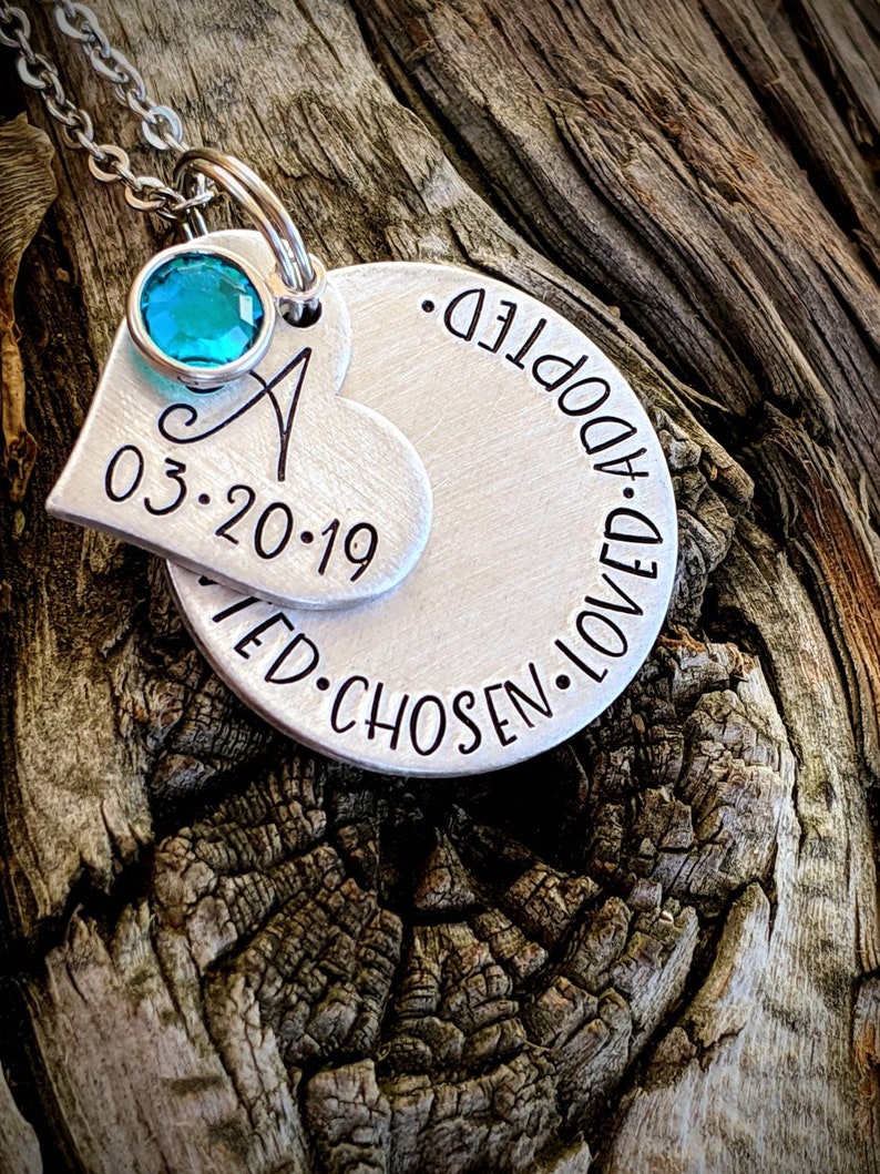 May include: Silver necklace with a heart charm that says "03.20.19" and a round charm that says "Adopted. Loved. Chosen." The heart charm has a turquoise gemstone.