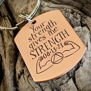May include: A copper-colored rectangular keychain with a silver ring. The keychain has the engraved text "Your strength gives me STRENGTH 08-13-21" with illustrations of flexing arms and a triangle symbol.