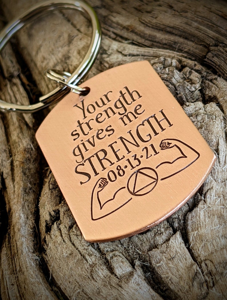 May include: A copper-colored rectangular keychain with a silver ring. The keychain has the engraved text "Your strength gives me STRENGTH 08-13-21" with illustrations of flexing arms and a triangle symbol.