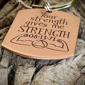 May include: A copper-colored keychain with the engraved text "Your strength gives me STRENGTH 08-13-21". The design includes illustrations of flexing arms and a triangle within a circle.