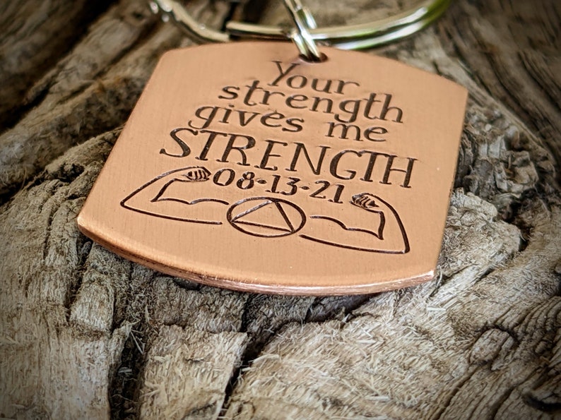 May include: A copper-colored keychain with the engraved text "Your strength gives me STRENGTH 08-13-21". The design includes illustrations of flexing arms and a triangle within a circle.