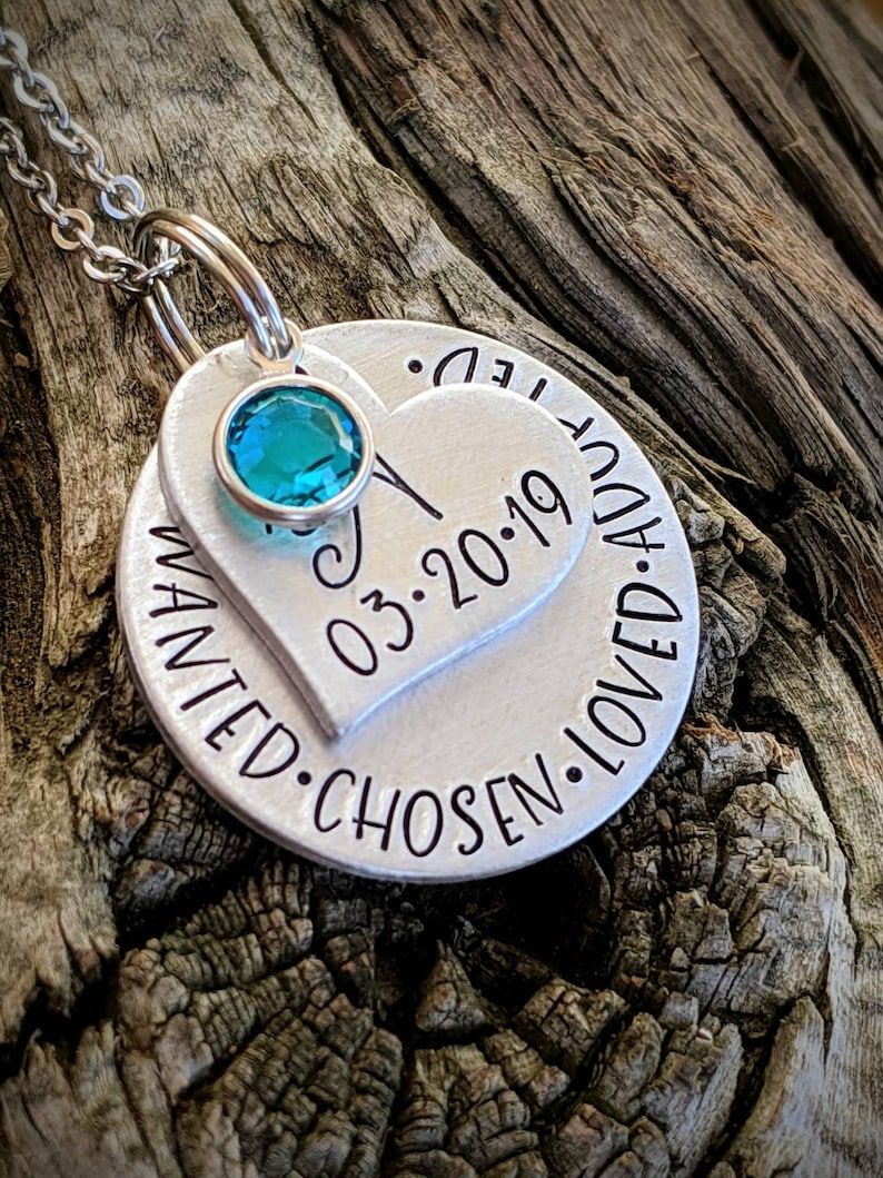 May include: Silver necklace with a heart-shaped pendant that says "Wanted, Chosen, Loved, Adored" and includes the date "03.2019". A turquoise gemstone is set in the center of the heart.