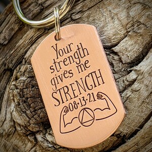 May include: A copper-colored dog tag keychain with the engraved text "Your strength gives me STRENGTH 08-13-21". The tag features illustrations of flexing arms and a circle with a triangle inside.