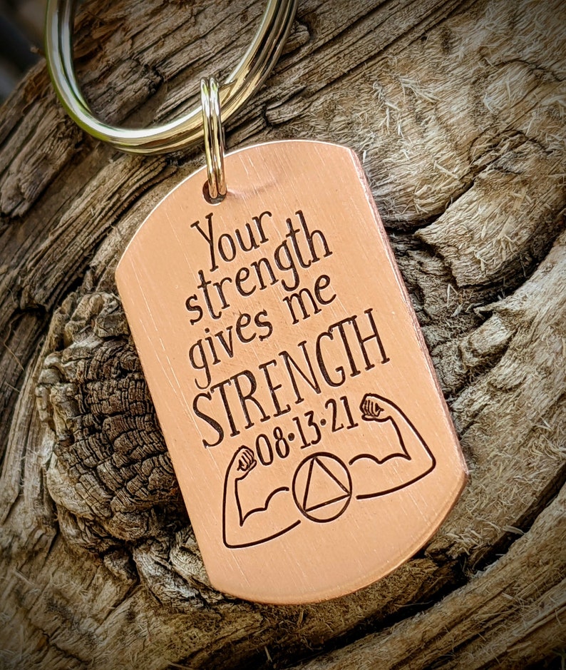 May include: A copper-colored dog tag keychain with the engraved text "Your strength gives me STRENGTH 08-13-21". The tag features illustrations of flexing arms and a circle with a triangle inside.