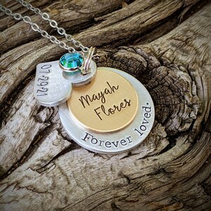 May include: A silver pendant necklace with multiple charms. The necklace features a heart-shaped charm with the date "07.29.21", a gold-colored charm with the name "Mayah Flores", and a silver charm with the words "Forever loved." A teal-colored gemstone is set above the charms.