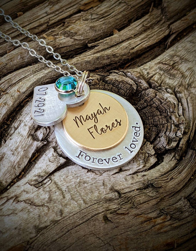 May include: A silver pendant necklace with multiple charms. The necklace features a heart-shaped charm with the date "07.29.21", a gold-colored charm with the name "Mayah Flores", and a silver charm with the words "Forever loved." A teal-colored gemstone is set above the charms.
