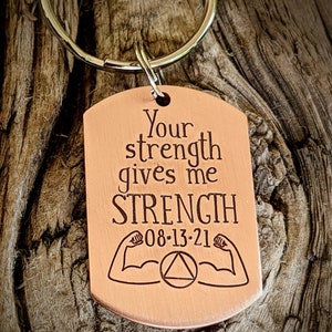 May include: A copper-colored dog tag keychain with the words "Your strength gives me STRENGTH" engraved in a bold font. Below the text are two flexing arm illustrations, the date 08-13-21, and a circle with a triangle inside.