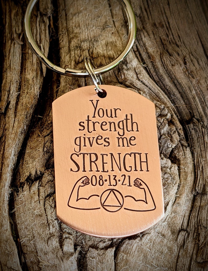 May include: A copper-colored dog tag keychain with the words "Your strength gives me STRENGTH" engraved in a bold font. Below the text are two flexing arm illustrations, the date 08-13-21, and a circle with a triangle inside.