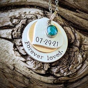 May include: Silver pendant necklace with a teal gemstone and a layered design. The pendant features the date "07.29.21" and the words "Forever loved." The necklace is displayed on a textured wooden surface.