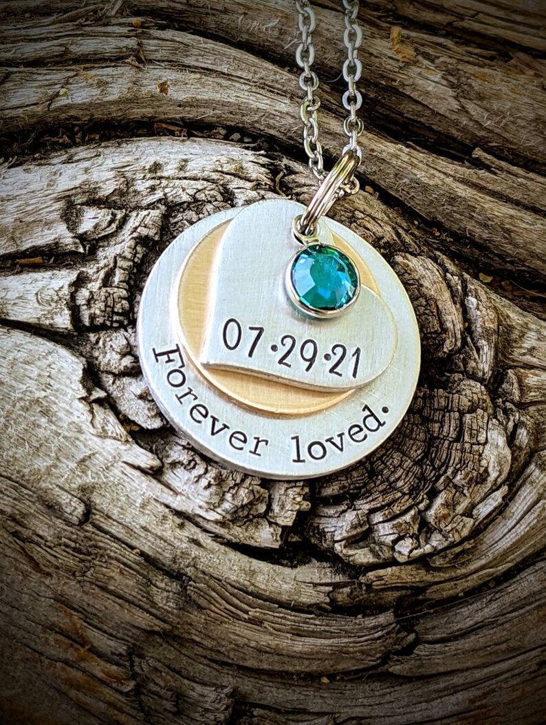 May include: Silver pendant necklace with a teal gemstone and a layered design. The pendant features the date "07.29.21" and the words "Forever loved." The necklace is displayed on a textured wooden surface.