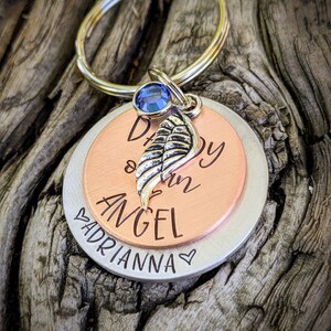 May include: A silver and copper keychain with a blue gem and a small silver wing charm. The keychain has the text "Daddy of an Angel" on the copper side and "Adrianna" on the silver side.