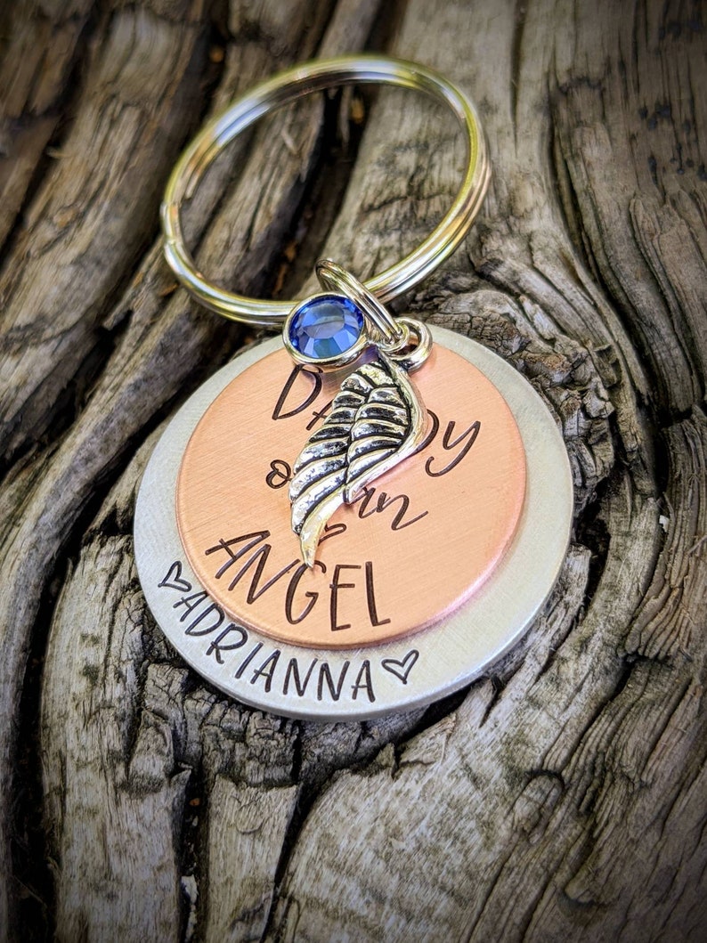 May include: A silver and copper keychain with a blue gem and a small silver wing charm. The keychain has the text "Daddy of an Angel" on the copper side and "Adrianna" on the silver side.
