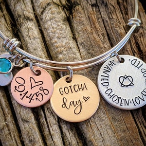 May include: Silver bangle bracelet with three charms. The charms are a heart with a turquoise stone, a copper charm with the date "03.14.58" and a heart, and a copper charm with the words "Gotcha Day" and a heart. The bracelet also has a silver charm with the words "Loved. Adopted. Chosen. Wanted." and a heart symbol.