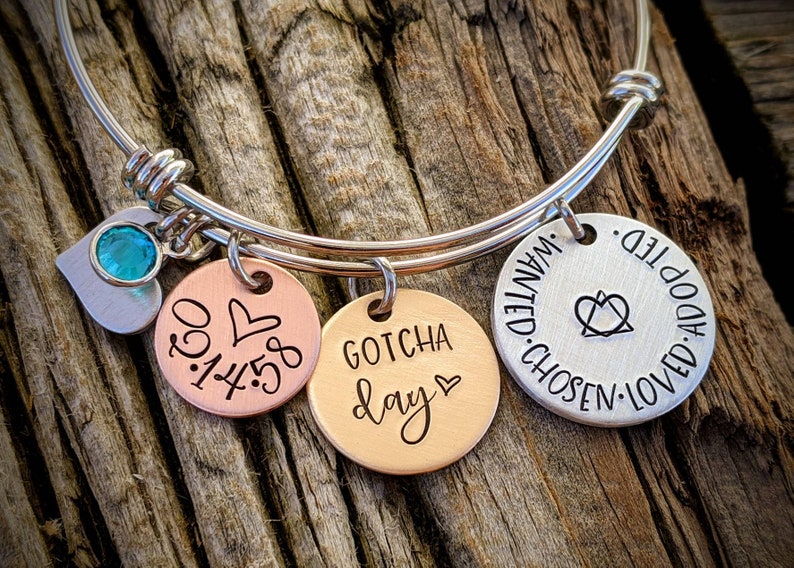 May include: Silver bangle bracelet with three charms. The charms are a heart with a turquoise stone, a copper charm with the date "03.14.58" and a heart, and a copper charm with the words "Gotcha Day" and a heart. The bracelet also has a silver charm with the words "Loved. Adopted. Chosen. Wanted." and a heart symbol.